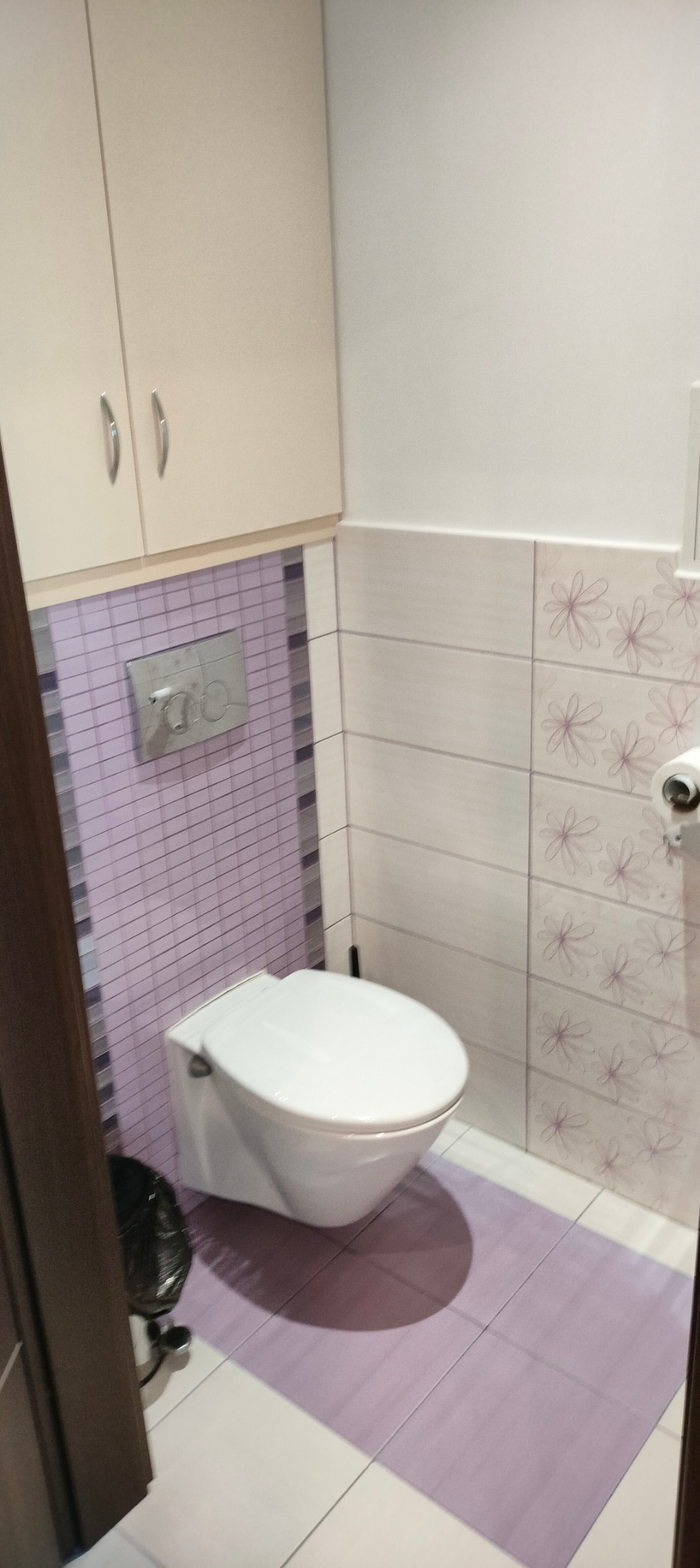 Wisząca toaleta w wykończonej łazience z fioletowymi akcentami na glazurze i podłodze, szafka nad toaletą, widoczny kosz na śmieci.