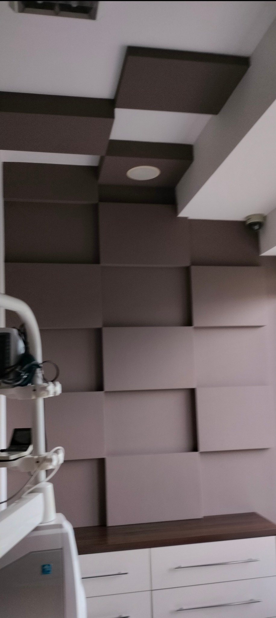 Nowoczesna ściana z paneli 3D w odcieniach szarości i brązu, oświetlenie punktowe w suficie, widoczny fragment wyposażenia gabinetu lekarskiego.