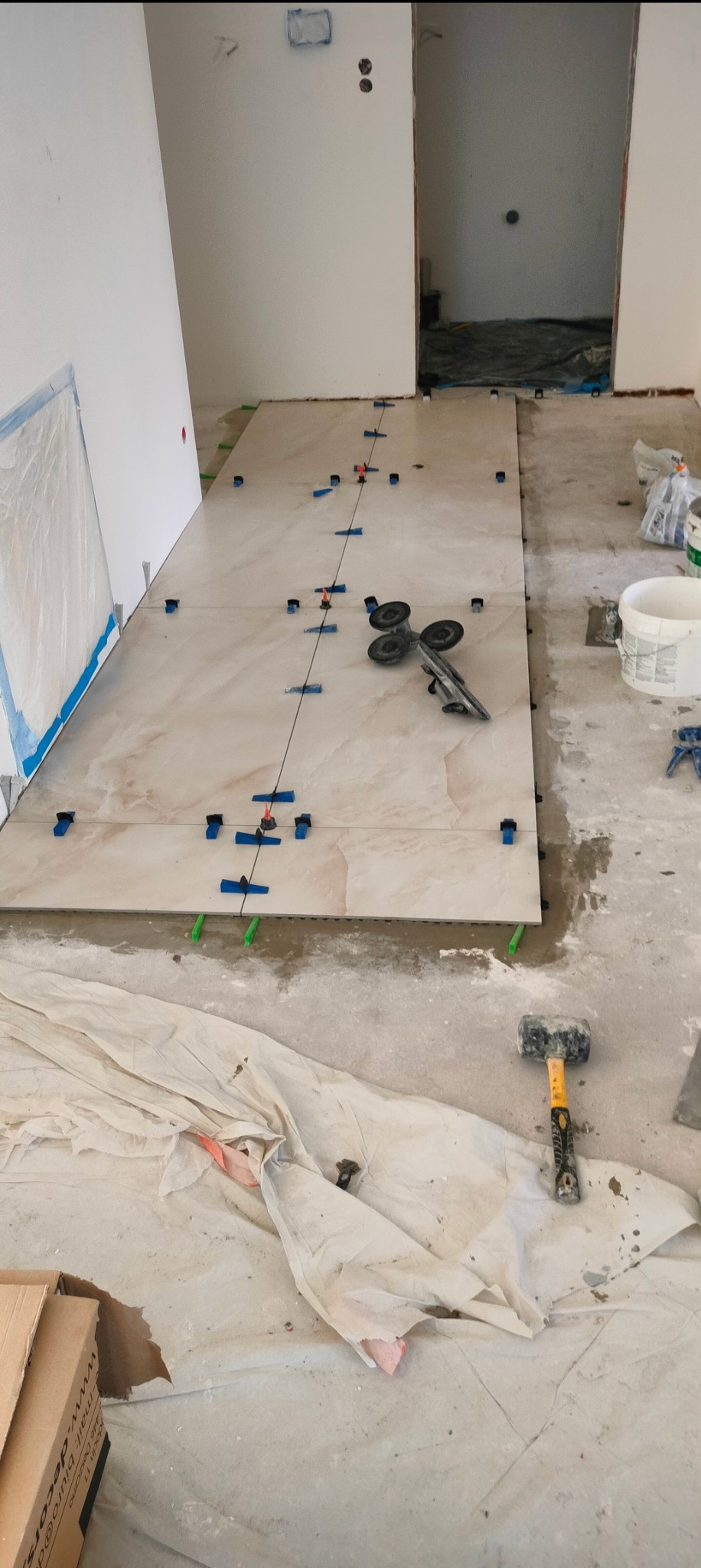 Układanie płytek podłogowych w trakcie remontu; duże beżowe płytki z widocznymi niebieskimi klipsami poziomującymi i przyrządem do cięcia płytek, sznurek wyznaczający linię prostą, w tle surowe...