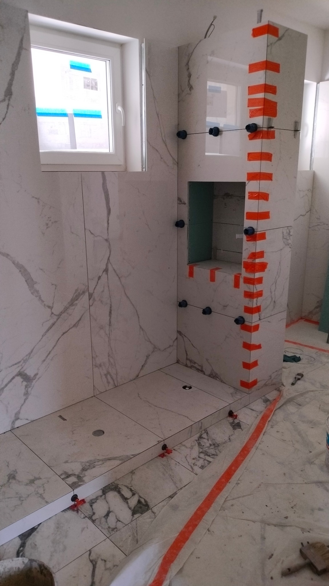 Łazienka w trakcie remontu: białe płytki imitujące marmur na ścianach i podłodze, poziomowanie płytek za pomocą klipsów i pomarańczowej taśmy, wnęka w ścianie wyłożona zielonym materiałem, okno w tle.