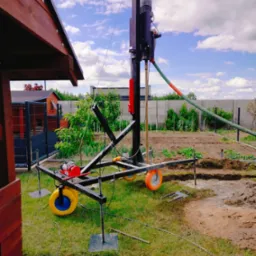 Wiertnica mobilna z pomarańczowymi kołami na trawie, przygotowana do wiercenia studni głębinowej na działce z widokiem na ogród i ogrodzenie.