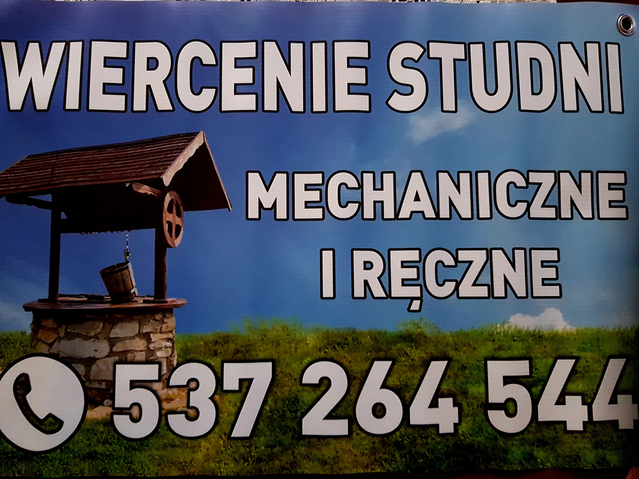 Baner reklamowy z grafiką studni, napisem 'Wiercenie Studni Mechaniczne i Ręczne' oraz numerem telefonu na tle błękitnego nieba i zielonej trawy.