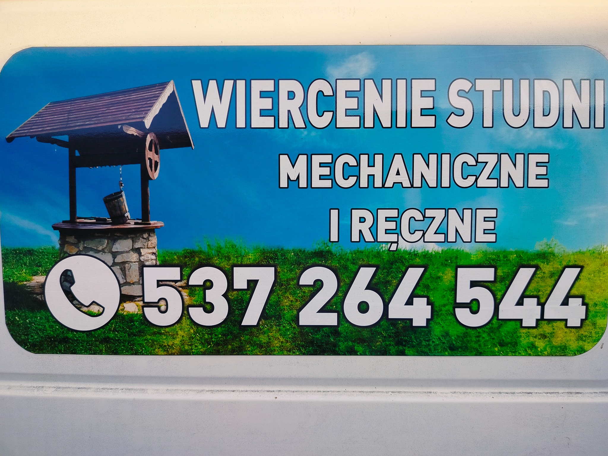 Reklama firmy w formie naklejki z rysunkiem studni z wiadrem, napisem 'Wiercenie Studni Mechaniczne i Ręczne' oraz numerem telefonu na tle błękitnego nieba i zielonej trawy.