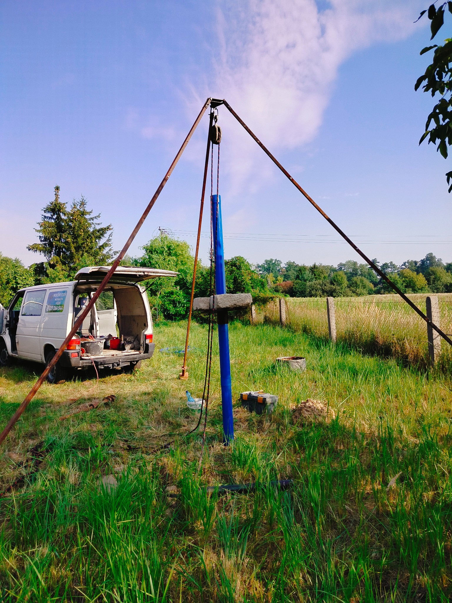 Wiercenie studni głębinowej na zewnątrz z użyciem wiertnicy linowej; na trawiastym terenie widoczny biały bus z otwartym bagażnikiem i narzędziami.