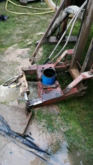 Fragment maszyny wiertniczej z niebieskim wiadrem w środku, otoczony błotem i trawą podczas prac wiertniczych.