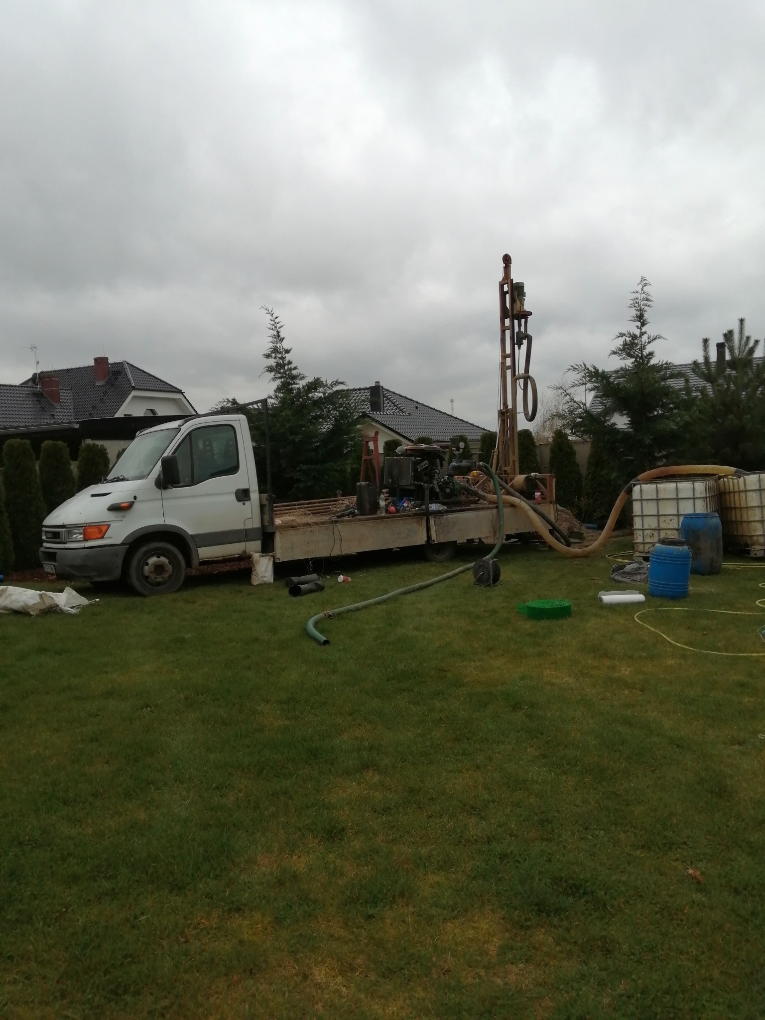 Biała ciężarówka z wiertnicą na platformie, przygotowana do wiercenia studni głębinowej na trawniku przed domem, widoczne zbiorniki na wodę i węże.