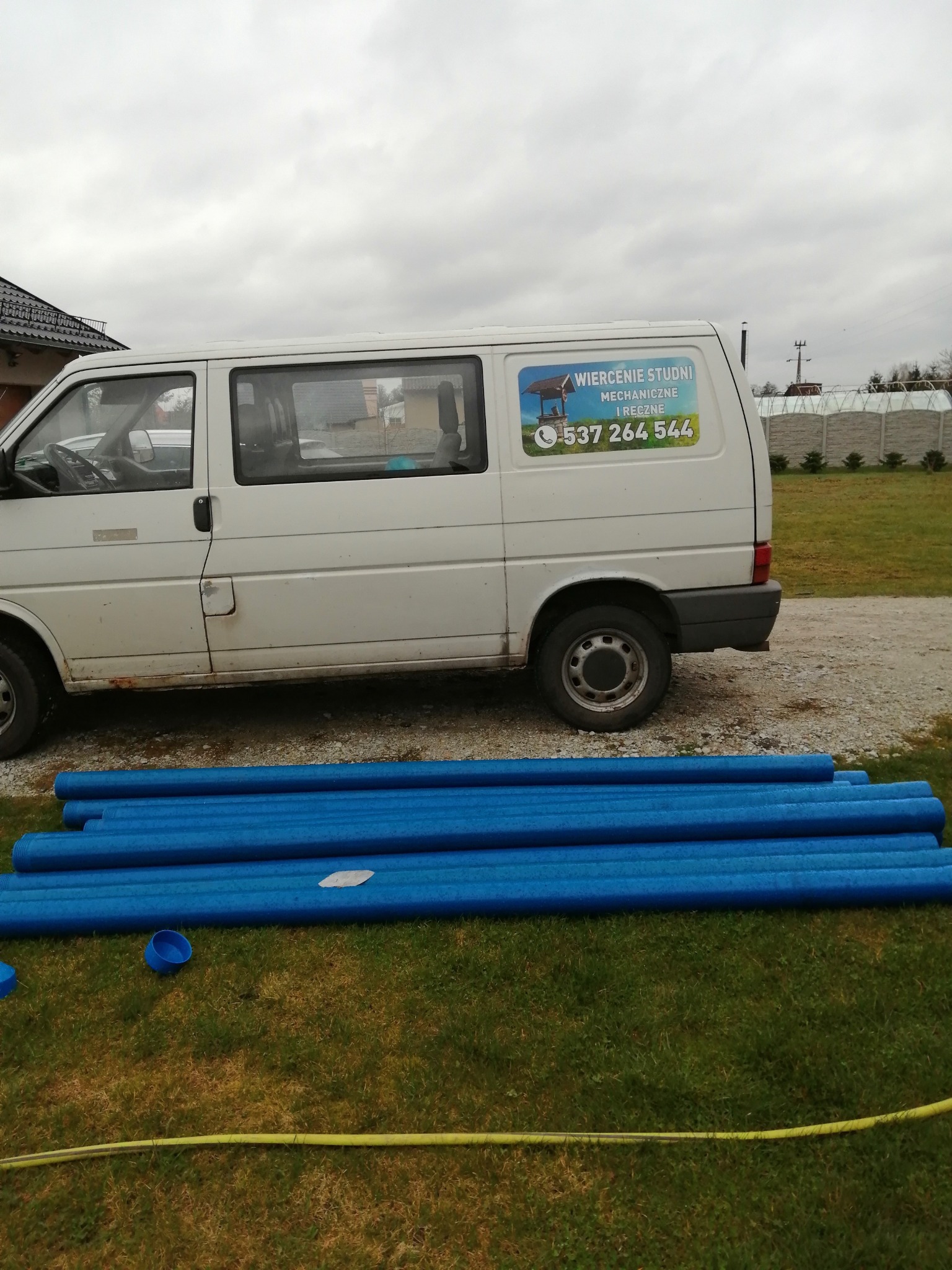 Biały bus z reklamą wiercenia studni, niebieskie rury na trawie, widok na dom i ogrodzenie w tle.