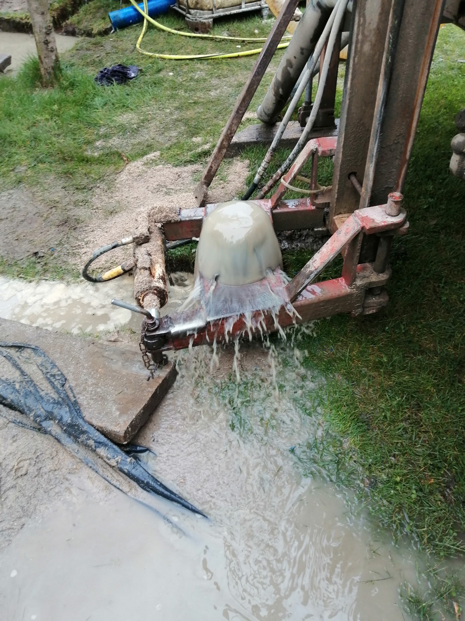 Wiercenie otworu w ziemi za pomocą wiertnicy, woda wypływa spod głowicy wiertniczej, błoto i trawa dookoła.