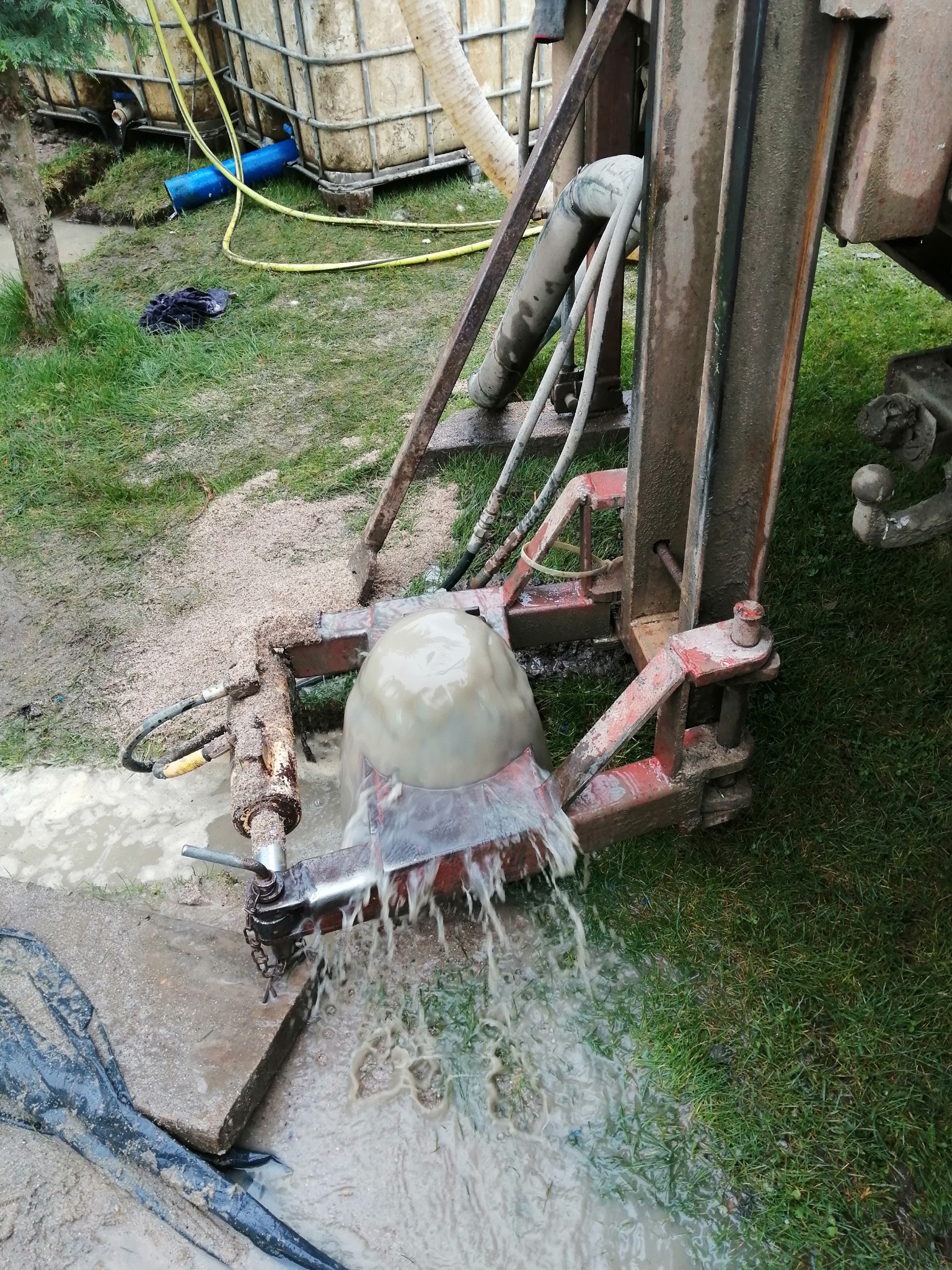 Proces wiercenia: woda wypływa spod mechanizmu wiertniczego na trawnik, widoczny zbiornik na wodę i błoto.