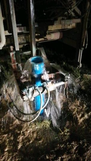 Wiercenie studni w nocy: woda tryska z niebieskiej rury, oświetlona przez reflektory wiertnicy, błotniste podłoże.