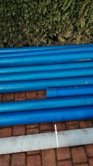 Stos niebieskich rur do wiercenia studni, ułożonych na czerwonej kostce brukowej, z zielonym żywopłotem w tle.