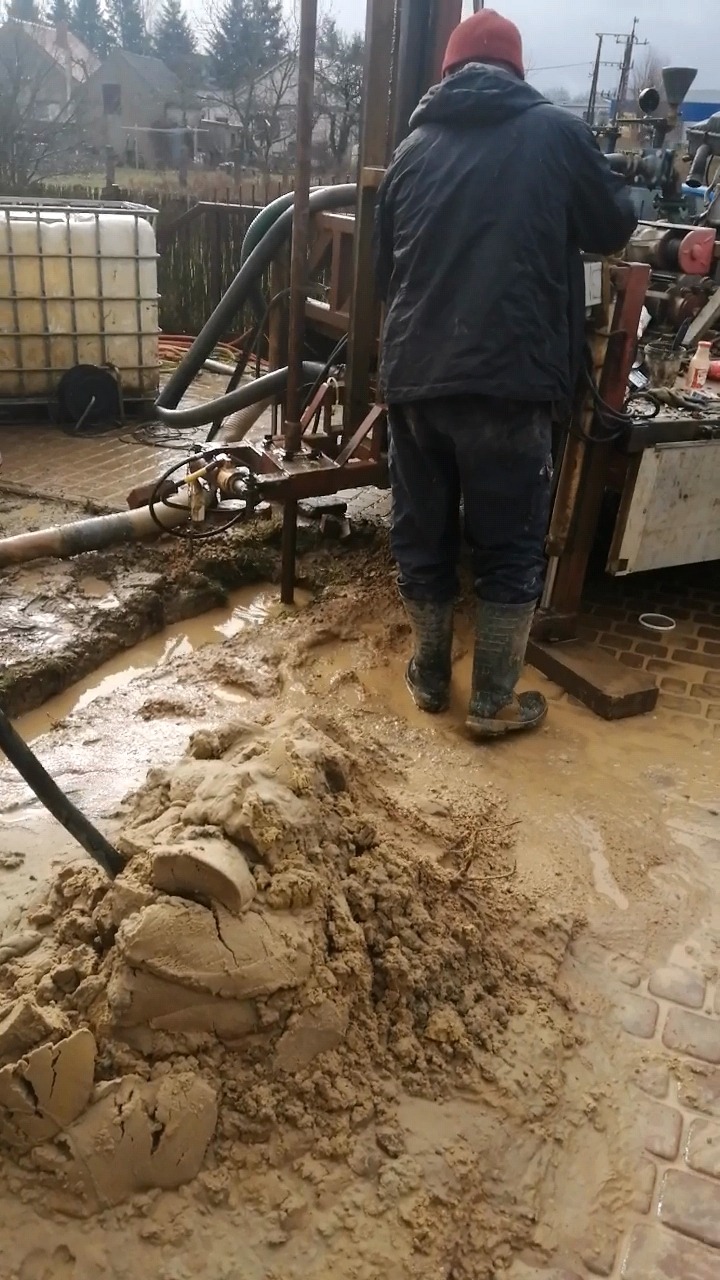 Pracownik w kaloszach obsługuje wiertnicę, wokół błoto i urobek ziemny, w tle biały zbiornik na wodę.
