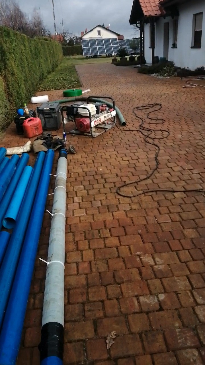 Przygotowane rury do wiercenia studni głębinowej leżące na brukowanym podjeździe, widoczna pompa do wody, narzędzia i budynek mieszkalny w tle.