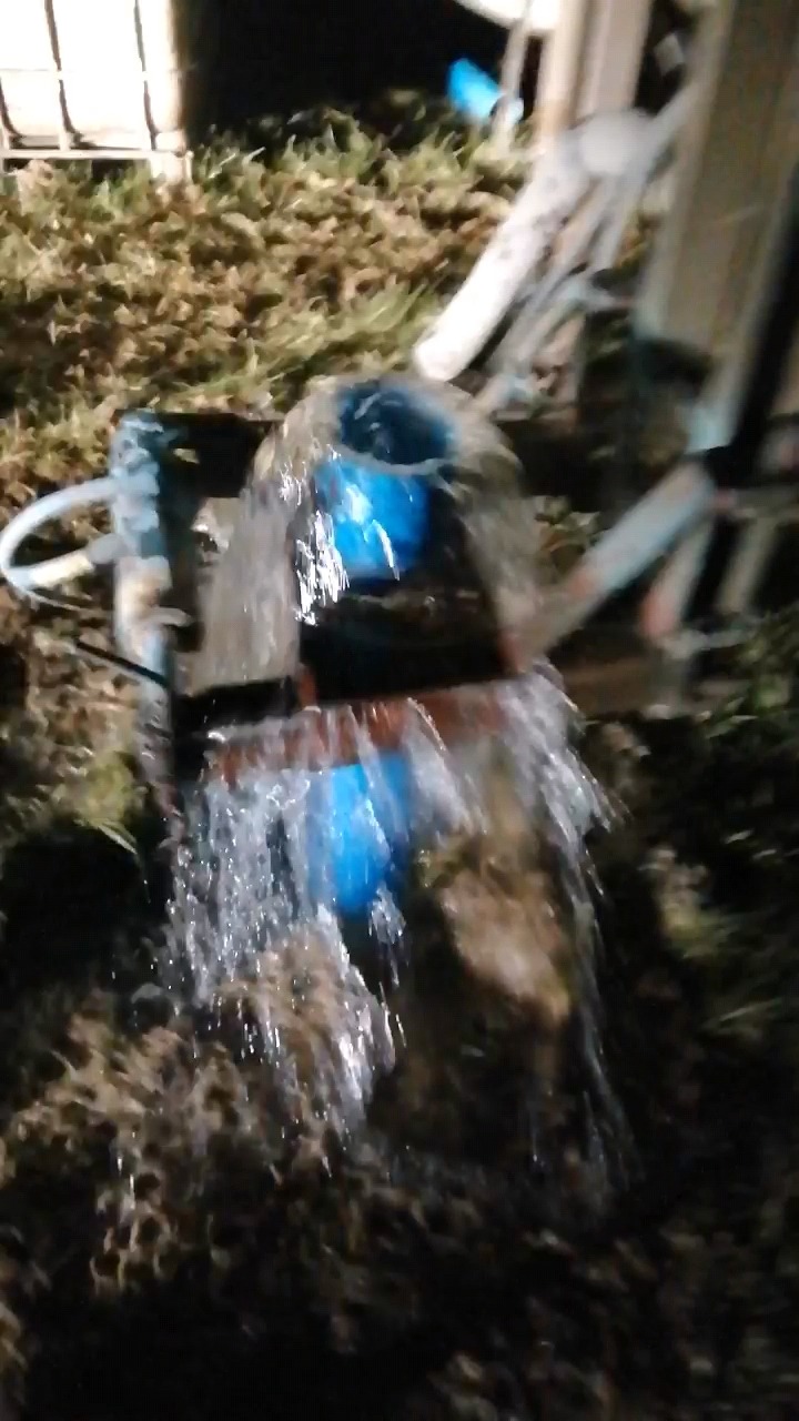 Woda wypływająca z rury studni głębinowej, widoczne elementy instalacji i otoczenie terenu w słabym oświetleniu nocnym.