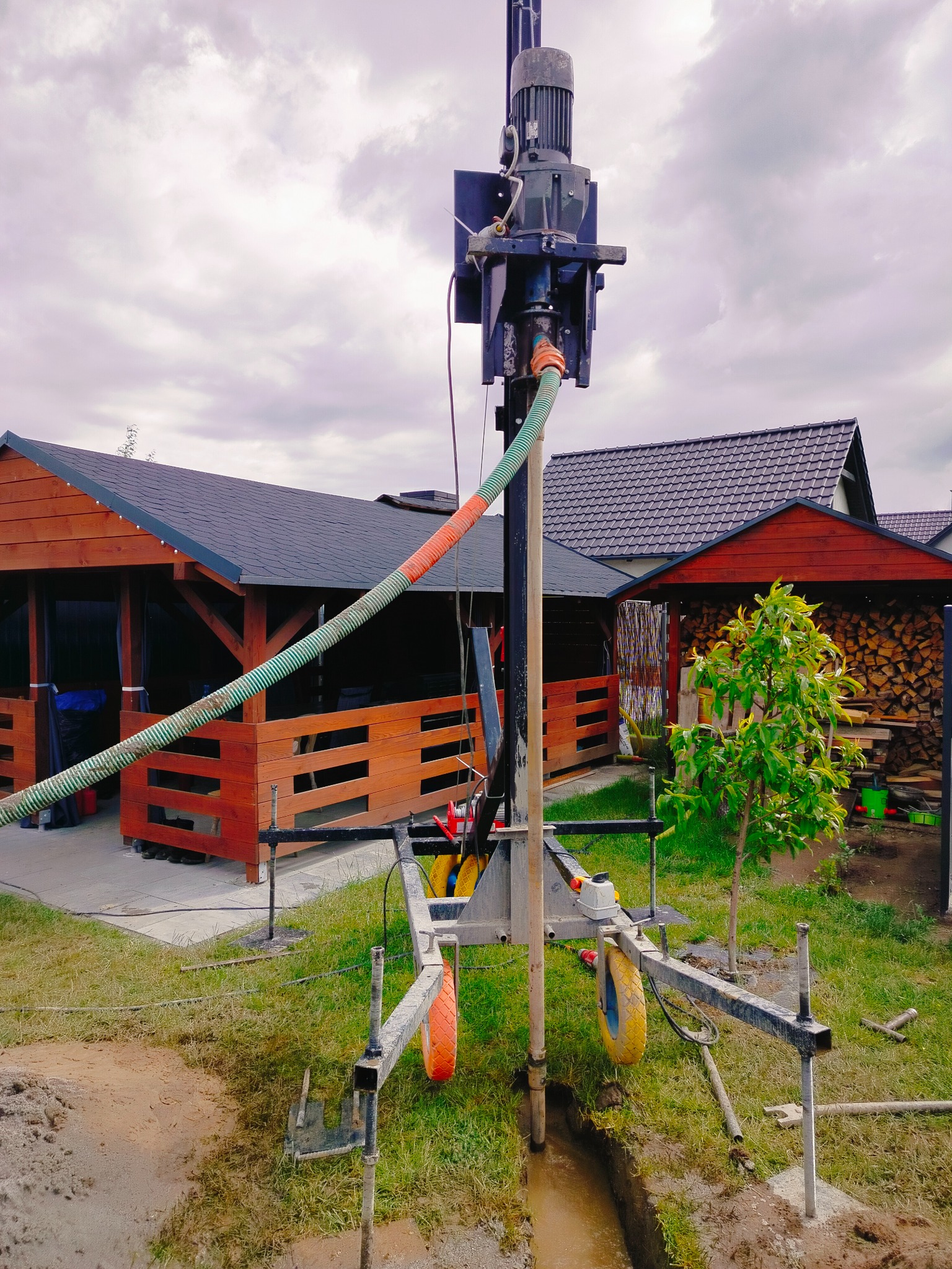 Wiertnica w ogrodzie, w trakcie pracy nad odwiertem. Widoczny fragment wykopu, fragment budynku w tle i pochmurne niebo.