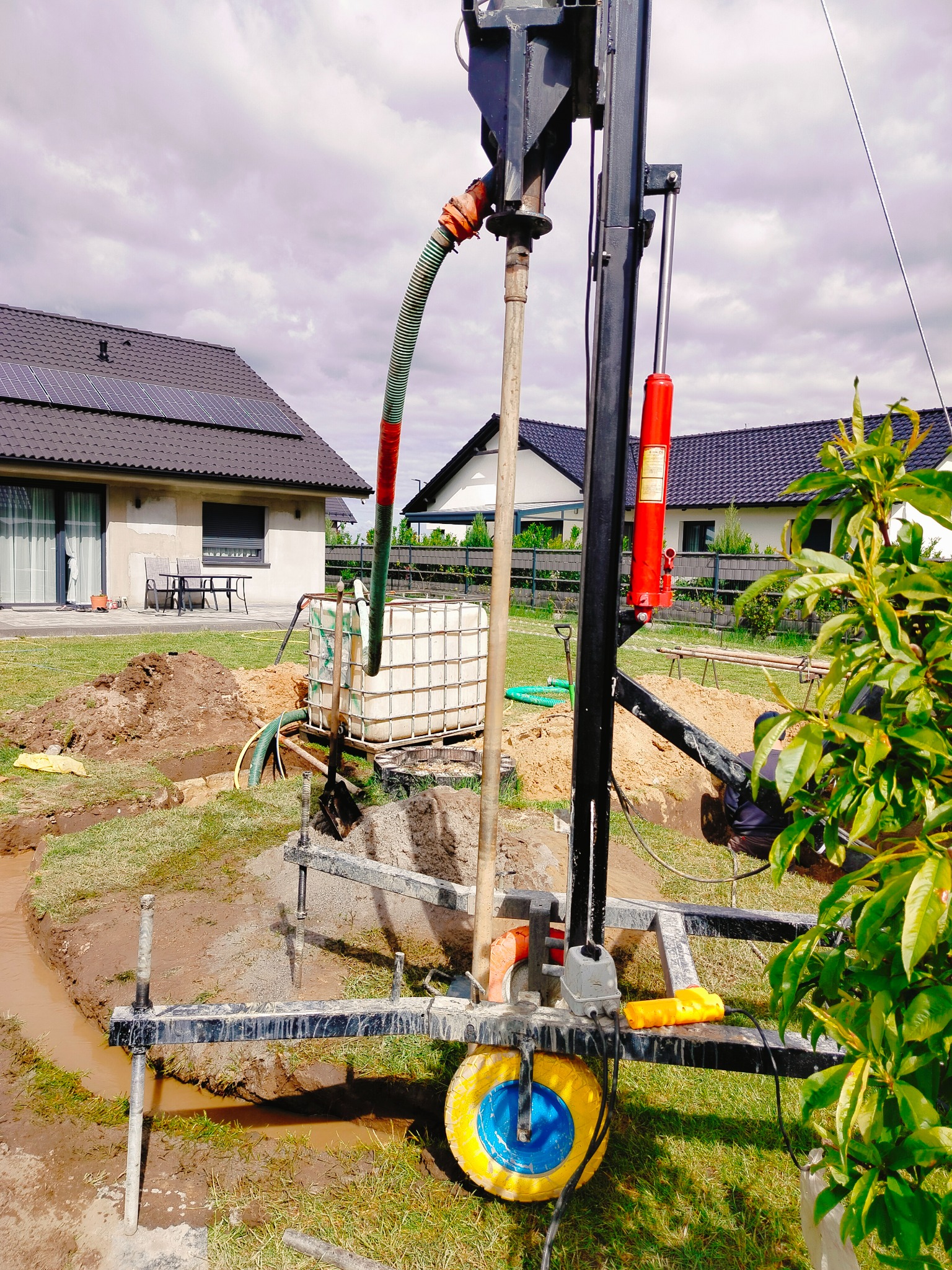Wiertnica do studni głębinowej w trakcie pracy na działce z domem. Widoczne wykopane rowy, zbiornik na wodę i elementy wiertnicy z żółtym kołem.