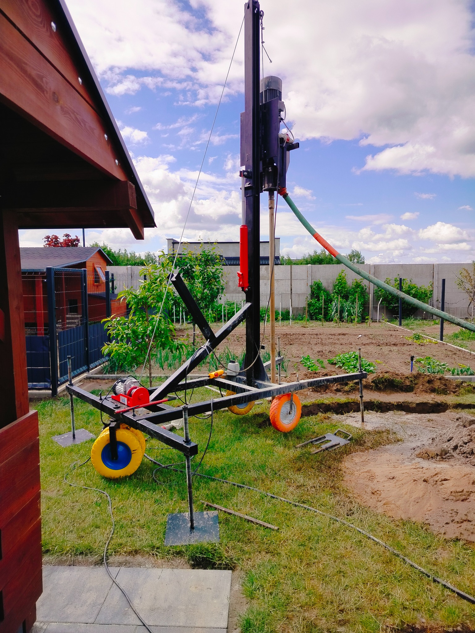 Wiertnica mobilna z pomarańczowymi kołami na trawie, przygotowana do wiercenia studni głębinowej na działce z widokiem na ogród i ogrodzenie.