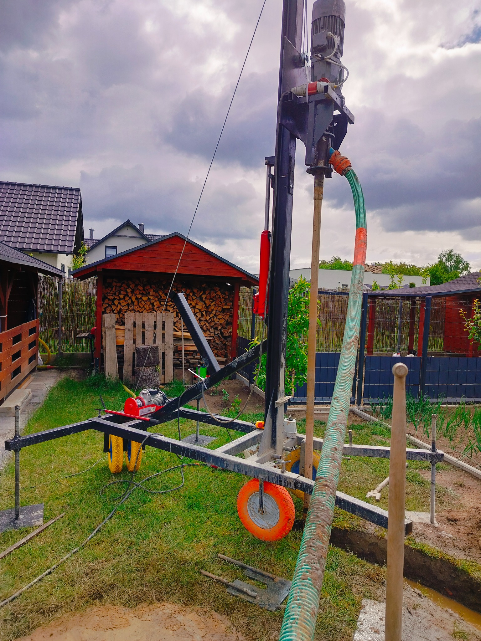 Wiertnica do studni głębinowej na trawniku, widoczna konstrukcja stalowa, silnik, przewód i narzędzia. W tle budynki mieszkalne i skład drewna.