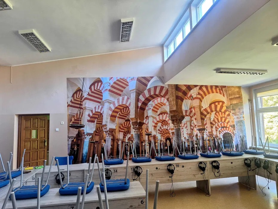 Ściana w sali lekcyjnej z fototapetą przedstawiającą wnętrze meczetu z charakterystycznymi łukami, poniżej rzędy krzeseł z niebieskimi siedziskami i słuchawkami, oświetlenie sufitowe.