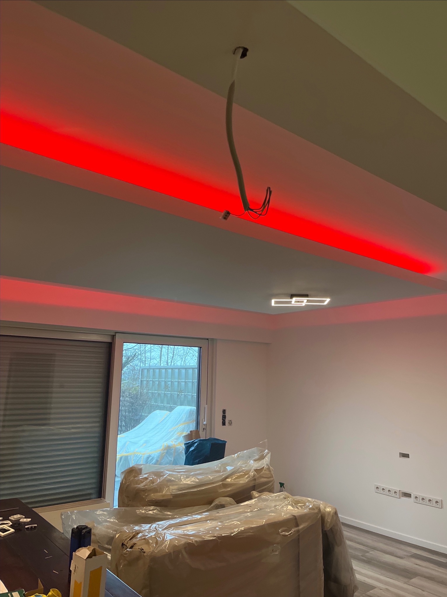 Wnętrze w trakcie remontu z zamontowanym oświetleniem LED w listwach sufitowych, widoczne kable, zakryte meble folią ochronną i gniazdka elektryczne w ścianie.