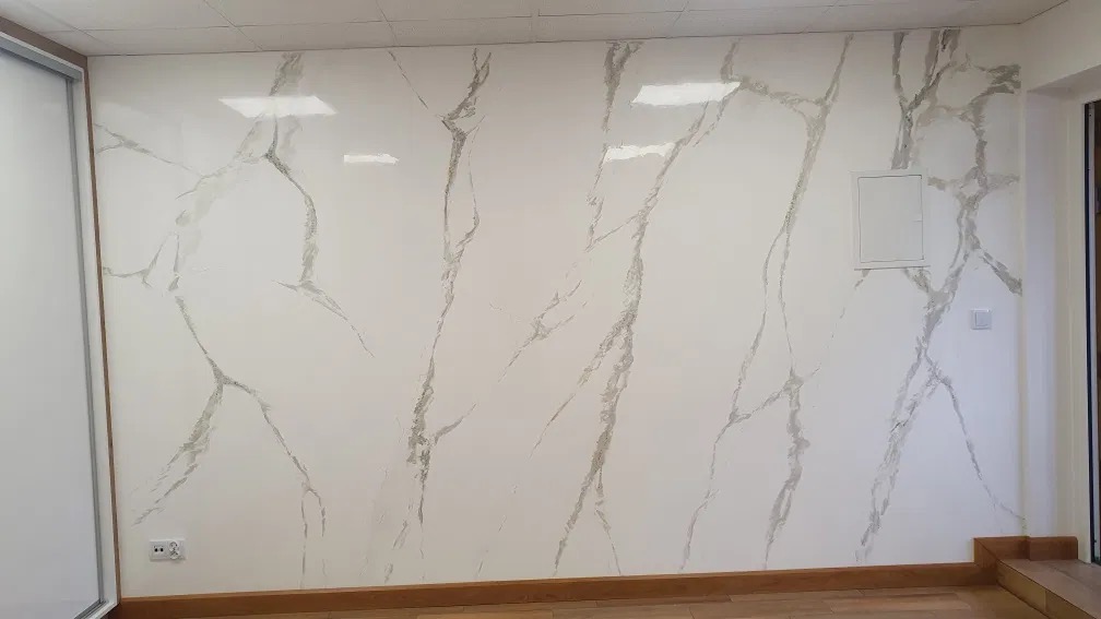 Ściana wykończona imitacją marmuru w odcieniach bieli i szarości, z widoczną skrzynką elektryczną i gniazdkiem, oświetlona światłem z sufitu.