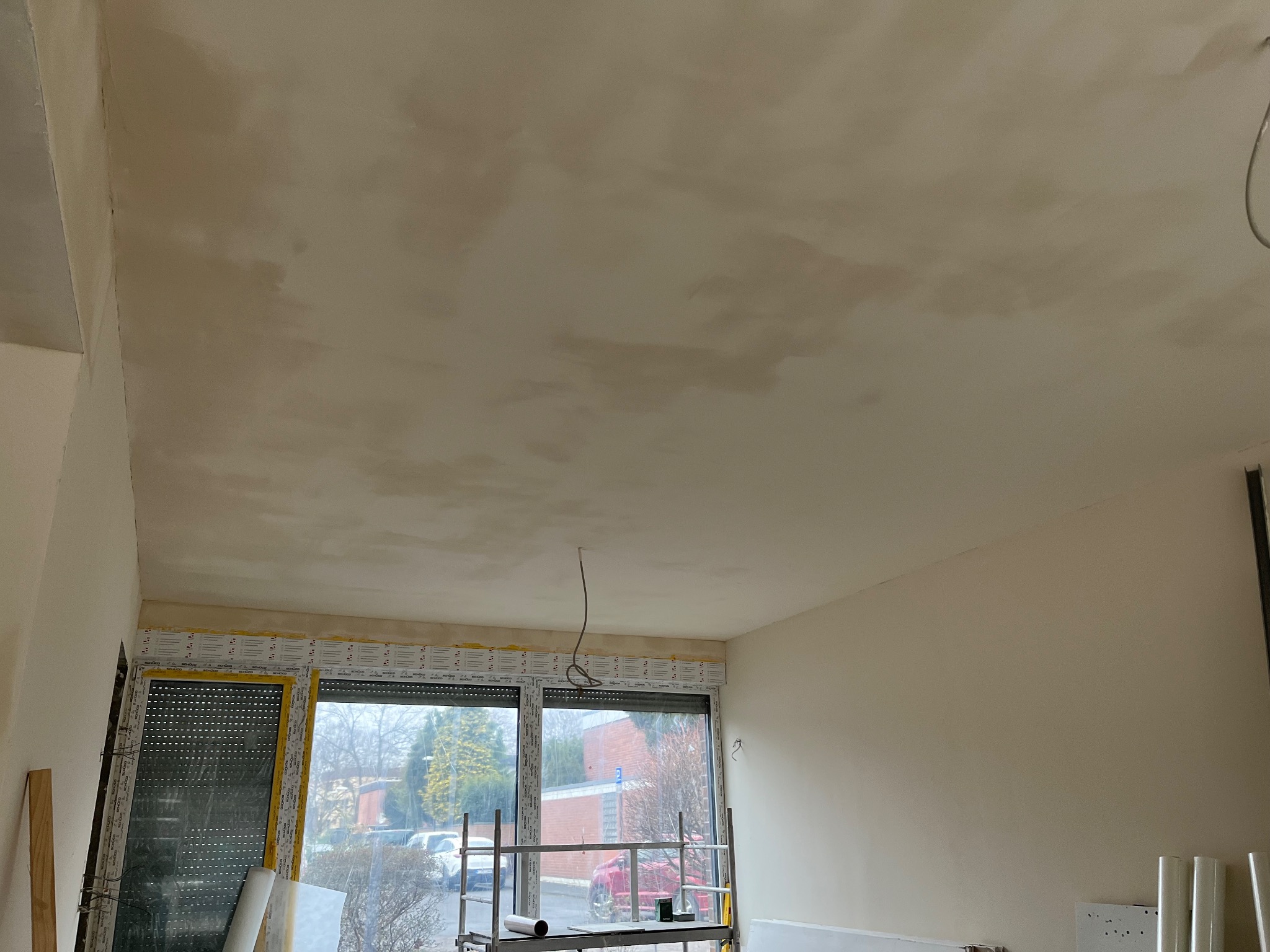 Nierównomiernie nałożona, świeża warstwa na suficie podczas prac wykończeniowych, widoczne ślady po szpachli, okno z roletami zabezpieczone folią ochronną, rusztowanie w pomieszczeniu.
