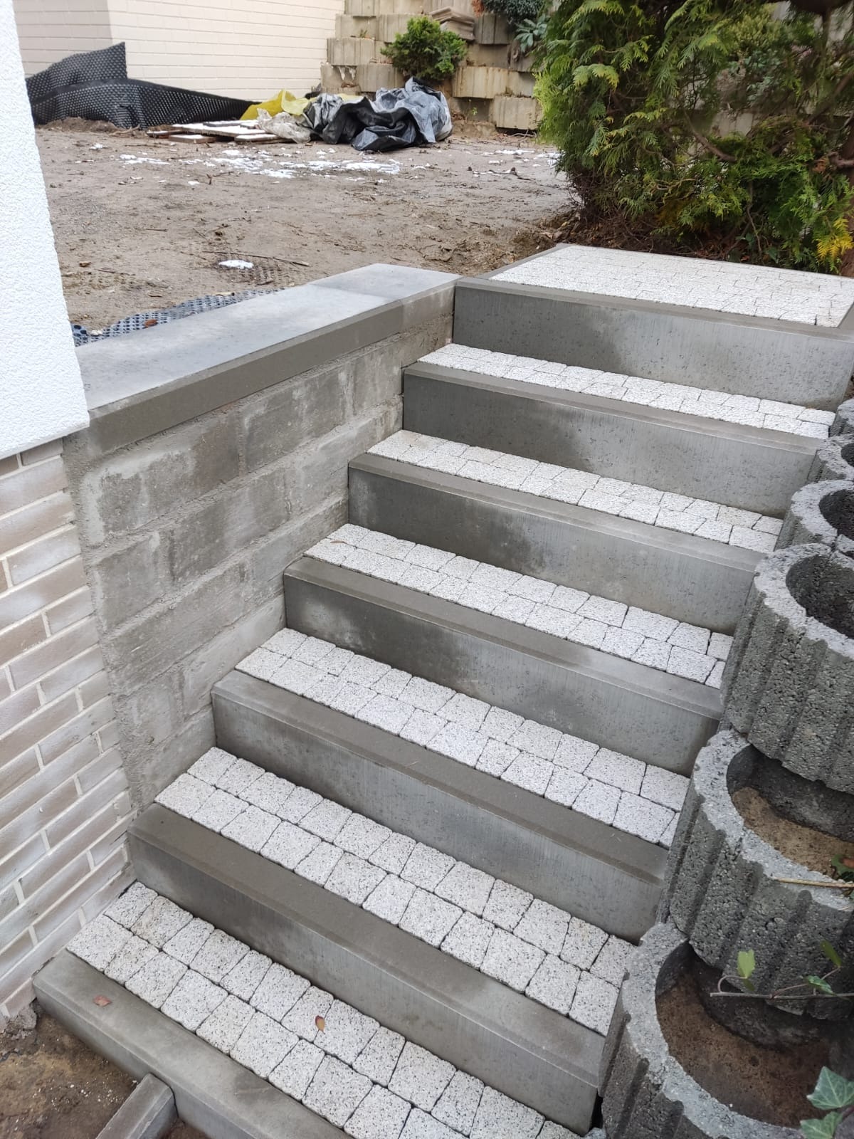 Betonowe schody zewnętrzne wykończone jasną kostką brukową, widoczne donice schodkowe po prawej stronie i fragment elewacji budynku po lewej.