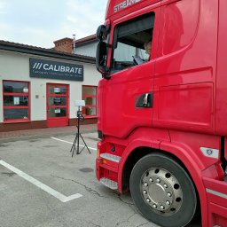 CALIBRATE - Czerwona ciężarówka Scania Streamline zaparkowana przed budynkiem serwisu tachografów Calibrate. Kierowca w kabinie wypełnia dokumenty, a na zewnątrz stoi urządzenie pomiarowe na statywie.