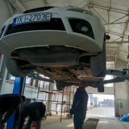 Biały samochód na podnośniku w warsztacie, widok od dołu, dwóch mechaników pracuje przy podwoziu, trzeci stoi obok, w tle brama garażowa i widok na ulicę.