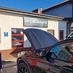 CALIBRATE - Czarny samochód z otwartą maską przed budynkiem serwisu, podłączony do niebieskiego urządzenia do obsługi klimatyzacji; w tle widoczny szyld 'CALIBRATE' oraz zaparkowany biały samochód dostawczy.