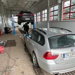CALIBRATE - Wnętrze warsztatu samochodowego z podnośnikiem: czarny SUV marki BMW na podnośniku, srebrne kombi BMW zaparkowane w pobliżu, widoczny pracownik warsztatu i narzędzia.