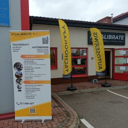CALIBRATE - Żółto-czarne flagi reklamowe i roll-up przed budynkiem warsztatu samochodowego oferującego serwis tachografów, klimatyzacji i inne usługi. Na roll-upie wypisane usługi i numer telefonu kontaktowego.