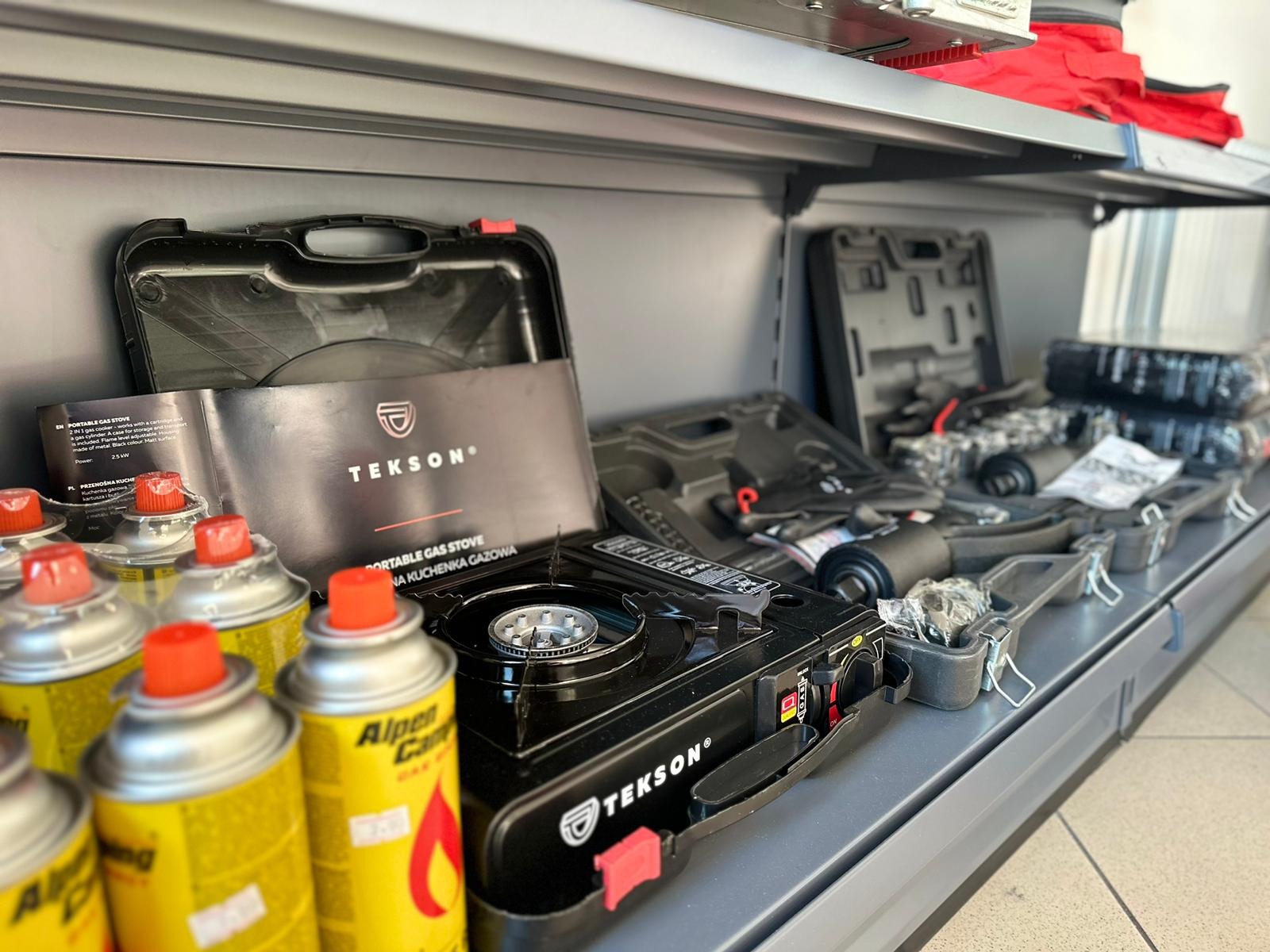 Półka sklepowa z przenośnymi kuchenkami gazowymi Tekson w czarnych walizkach i puszkami z gazem Alpen Camping, widok z boku.