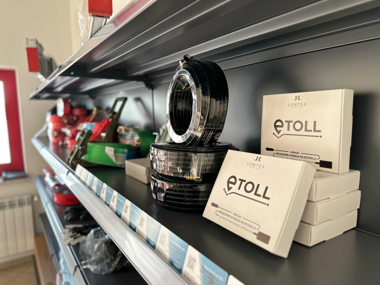 Półka z akcesoriami samochodowymi: zwoje czarnego kabla w folii oraz pudełka z urządzeniami e-TOLL firmy LONTEX, widoczne etykiety i napisy informacyjne.