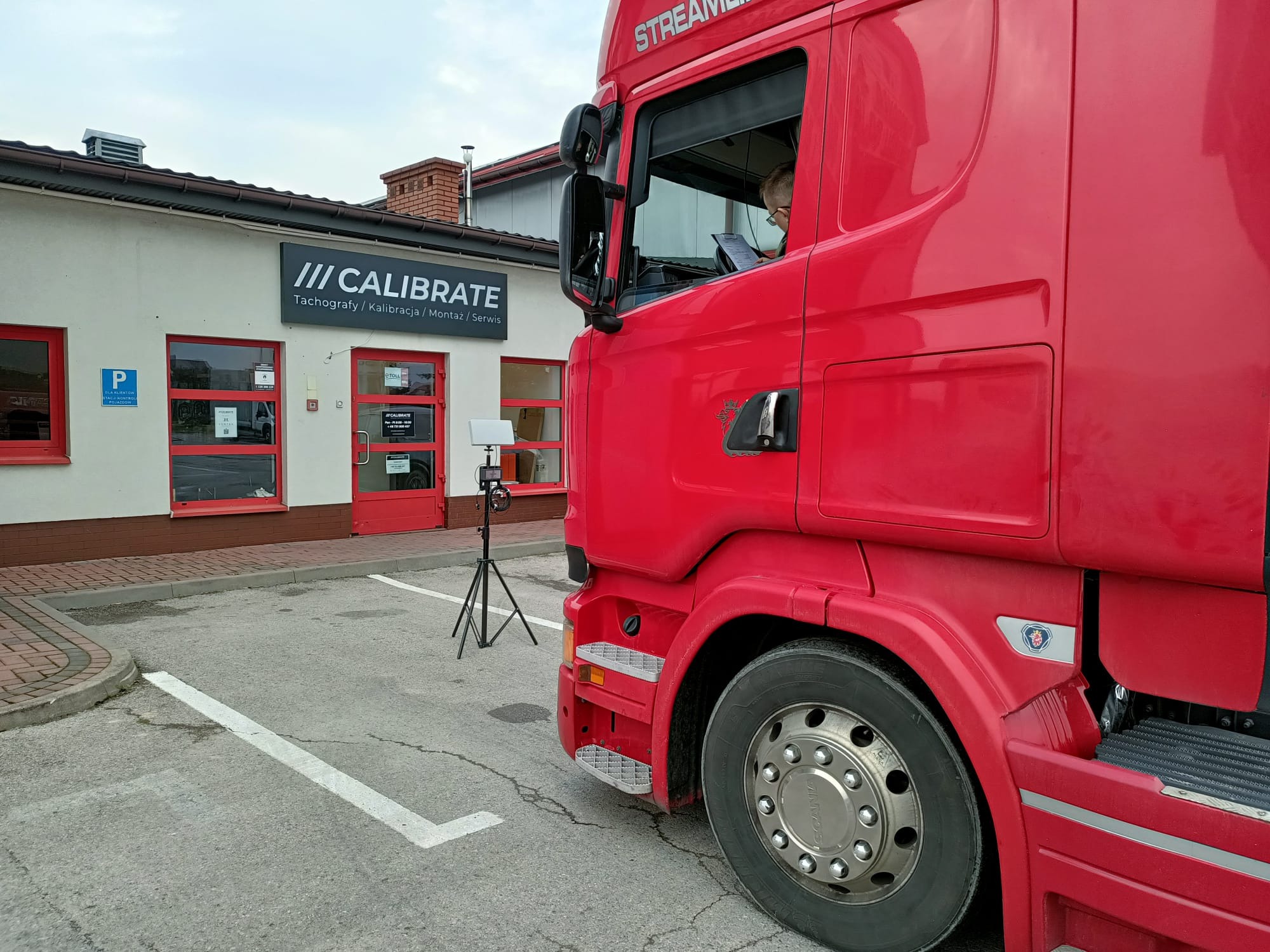 Czerwona ciężarówka Scania Streamline zaparkowana przed budynkiem serwisu tachografów Calibrate. Kierowca w kabinie wypełnia dokumenty, a na zewnątrz stoi urządzenie pomiarowe na statywie.
