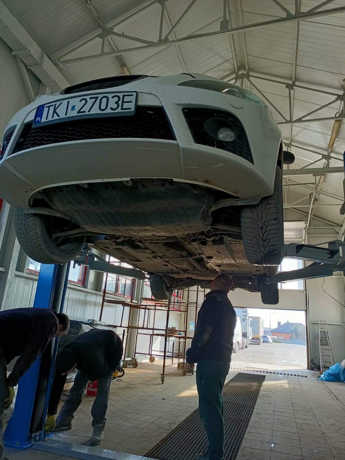 Biały samochód na podnośniku w warsztacie, widok od dołu, dwóch mechaników pracuje przy podwoziu, trzeci stoi obok, w tle brama garażowa i widok na ulicę.