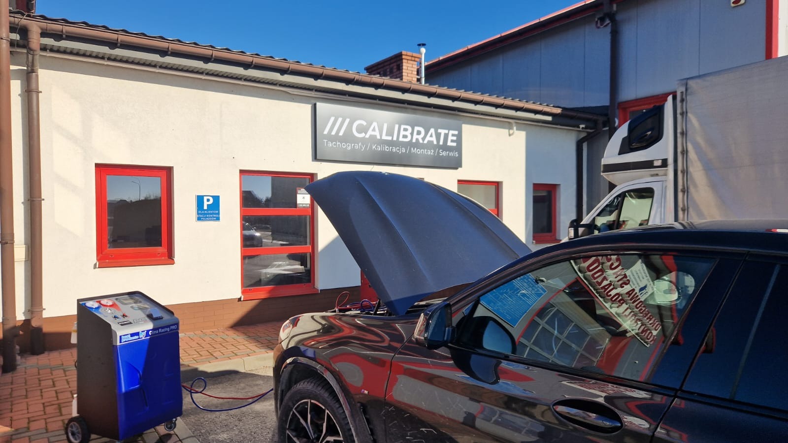 Czarny samochód z otwartą maską przed budynkiem serwisu, podłączony do niebieskiego urządzenia do obsługi klimatyzacji; w tle widoczny szyld 'CALIBRATE' oraz zaparkowany biały samochód dostawczy.
