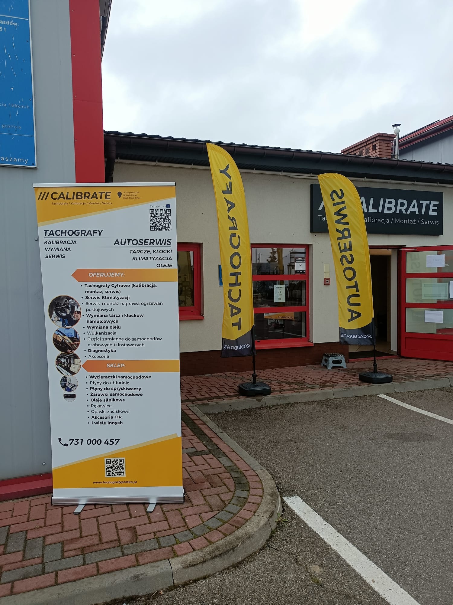 Żółto-czarne flagi reklamowe i roll-up przed budynkiem warsztatu samochodowego oferującego serwis tachografów, klimatyzacji i inne usługi. Na roll-upie wypisane usługi i numer telefonu kontaktowego.