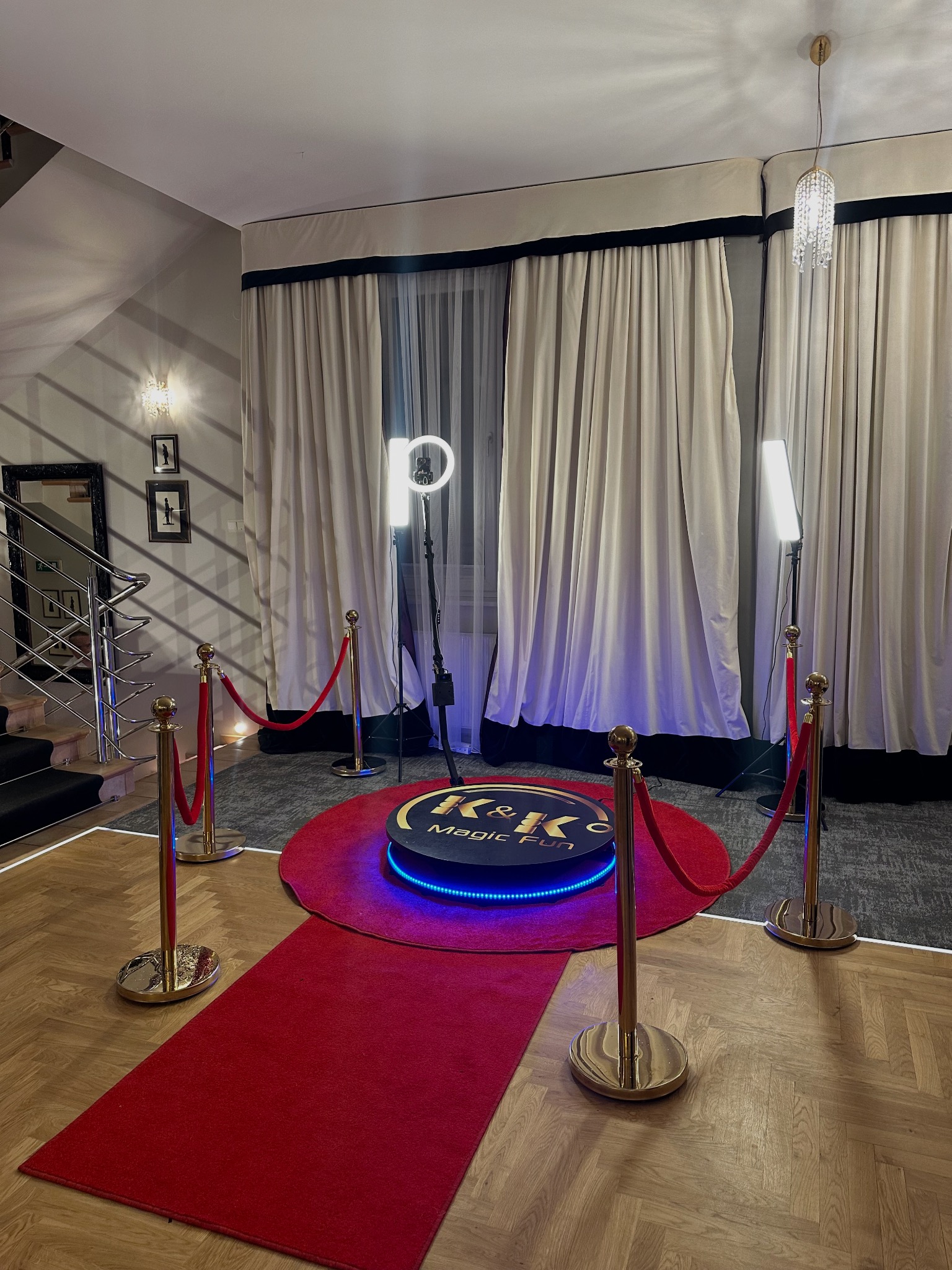 Elegancka fotobudka 360 z oświetleniem LED i czerwonym dywanem, otoczona złotymi słupkami z czerwonym sznurem w przestronnym wnętrzu z dekoracyjnymi zasłonami.
