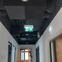 Marmil - Korytarz z czarnym sufitem, na którym widoczne są rury, przewody, lampy i znak ewakuacyjny. Na środku drewniana drabina, z wkrętarką na jednym ze szczebli, w tle drzwi. Na podłodze leży radio...