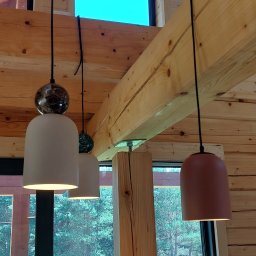 Marmil - Wnętrze drewnianego domu z trzema wiszącymi lampami o różnej kolorystyce kloszy (biały, biały z marmurkową kulą, terakota) zamontowanymi na drewnianych belkach stropowych z widocznym oknem...