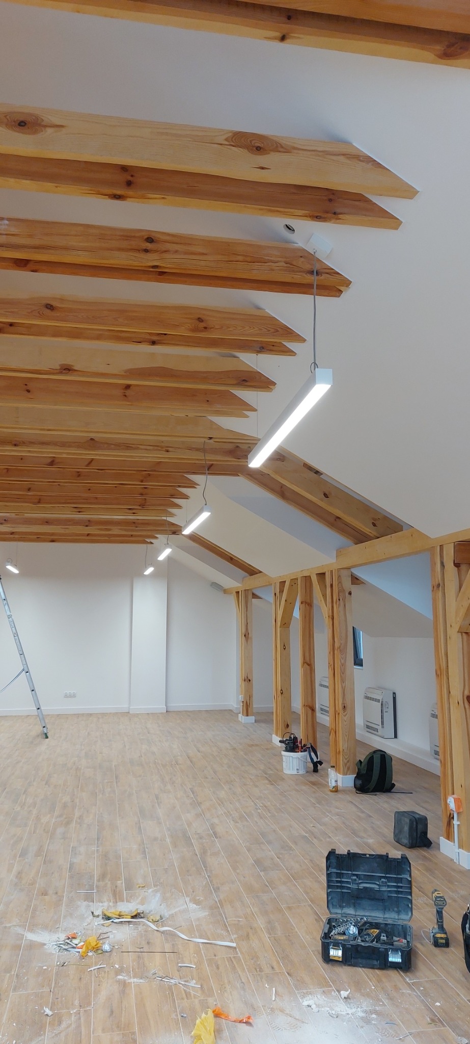 Wnętrze poddasza z drewnianą konstrukcją stropu i oświetleniem LED w trakcie montażu, widoczne narzędzia i pozostałości po pracach na podłodze.