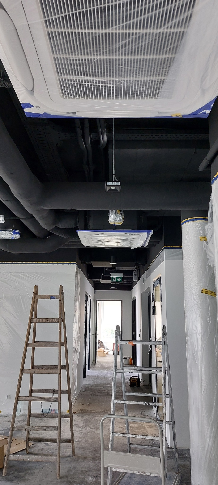 Sufit z instalacją klimatyzacji zabezpieczoną folią malarską, widoczne rury instalacyjne, czarny sufit, dwie drabiny w korytarzu podczas prac wykończeniowych.