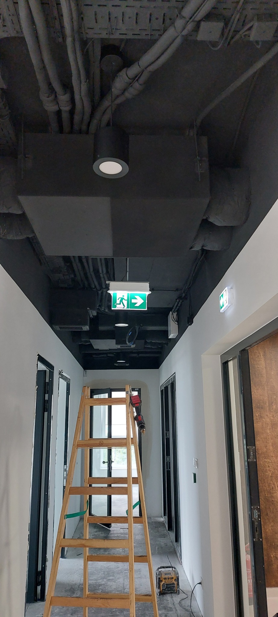Korytarz z czarnym sufitem, na którym widoczne są rury, przewody, lampy i znak ewakuacyjny. Na środku drewniana drabina, z wkrętarką na jednym ze szczebli, w tle drzwi. Na podłodze leży radio...