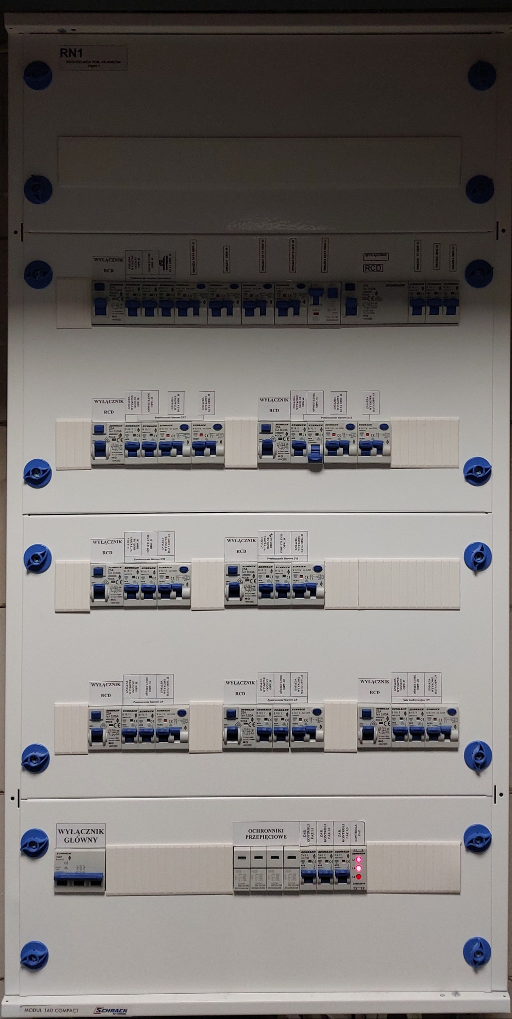 Rozdzielnica elektryczna z wyłącznikami różnicowoprądowymi, zabezpieczeniami przepięciowymi i wyłącznikiem głównym, widok z przodu na ścianie.