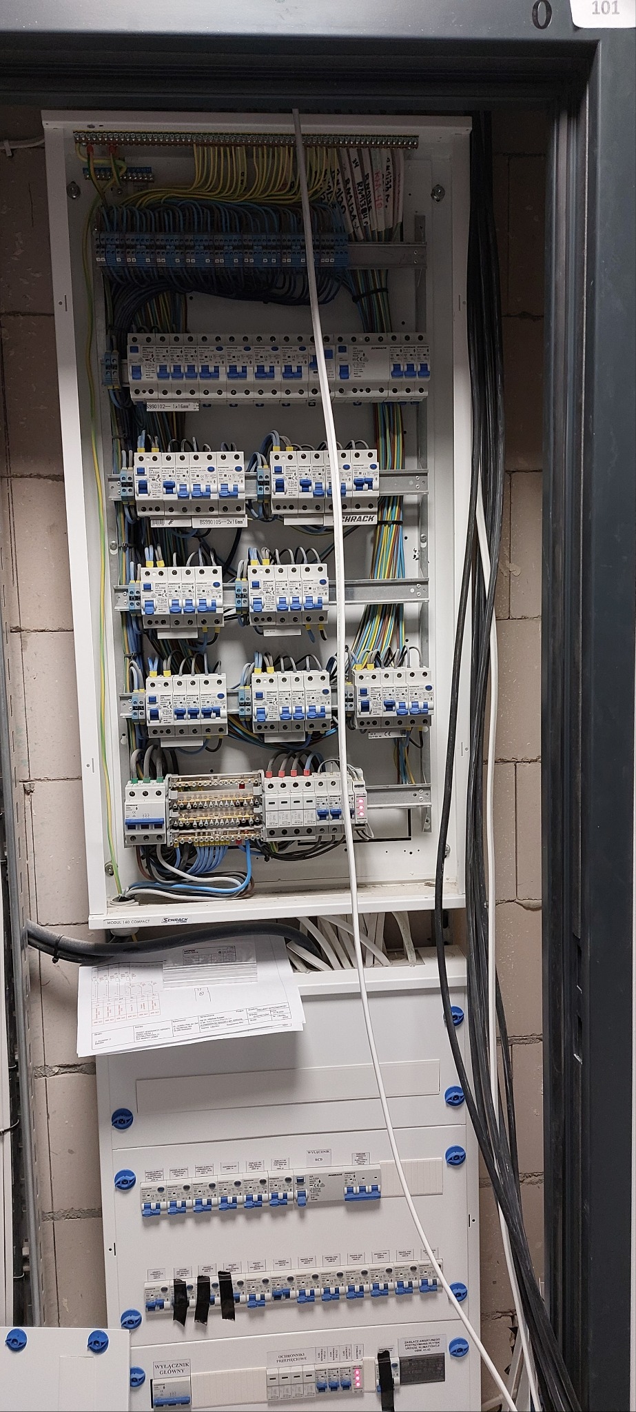 Rozdzielnica elektryczna w trakcie montażu, widoczne kable, bezpieczniki, listwy zaciskowe i schemat podłączeń, ściana z cegły w tle.