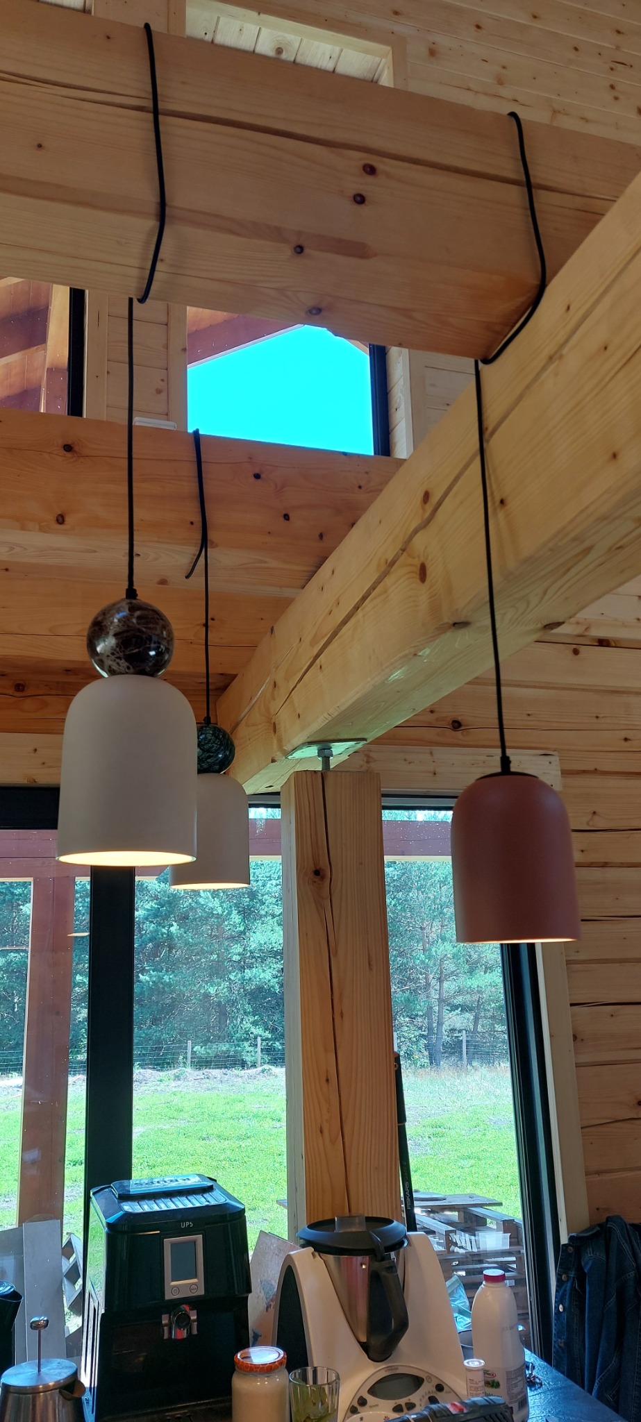 Wnętrze drewnianego domu z trzema wiszącymi lampami o różnej kolorystyce kloszy (biały, biały z marmurkową kulą, terakota) zamontowanymi na drewnianych belkach stropowych z widocznym oknem...