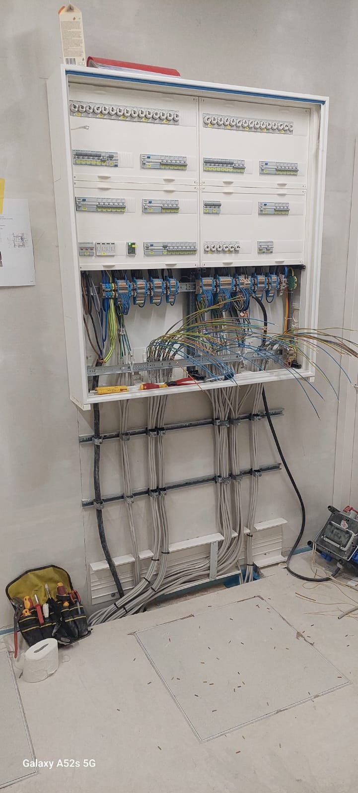Rozdzielnica elektryczna w trakcie montażu, widoczne liczne przewody, bezpieczniki, narzędzia w torbie obok, oraz otwarte klapy w podłodze.