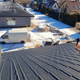 Nowy, grafitowy dach z blachy trapezowej w trakcie montażu, widok z góry na częściowo pokryty dach, w tle biały samochód dostawczy i zasypana śniegiem ulica z zabudowaniami.