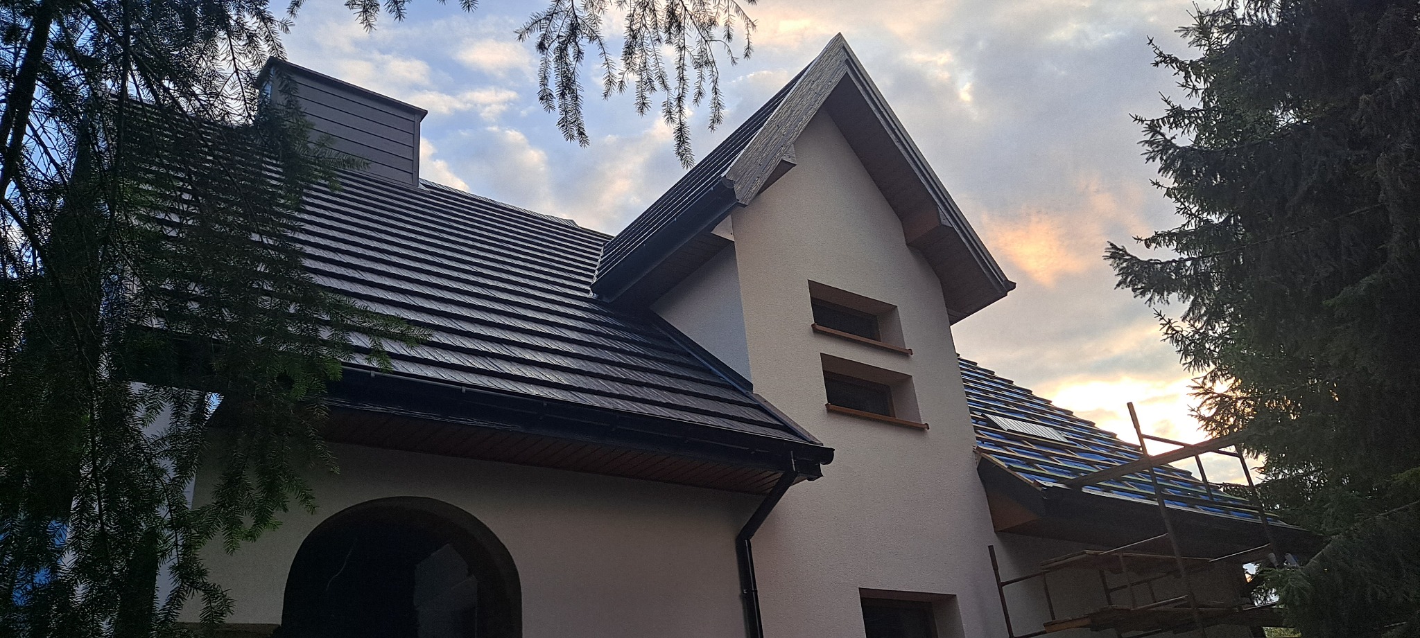 Nowoczesny dom z ciemną dachówką ceramiczną, widoczne obróbki blacharskie i fragment rusztowania podczas prac dekarskich, na tle zachodzącego słońca.